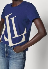 Osoba v tmavě modrém tričku s velkými bílými překrývajícími se písmeny a světle modrých džínách, ruka v kapse, proti neutrálnímu pozadí.