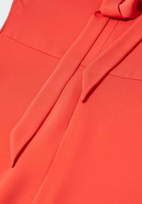 Korallefarbene Bluse aus glattem Stoff, mit einem Halstuch und gerader Schnittführung, leicht glänzend und mit minimalen Nähdetails.