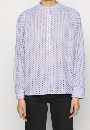 Persona con una blusa lavanda de manga larga, abotonada, con bordado de ojales y pantalones negros, de pie frente a un fondo blanco liso.