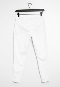 Bílé úzké kalhoty vyrobené z bavlněné směsi, s hladkou texturou, dvěma zadními kapsami a pasem s poutky na opasek, zavěšené na černém háčku.