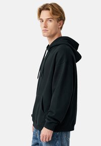 Felpa con cappuccio nera in tessuto morbido, con tasca a marsupio, cappuccio con coulisse e polsini a costine, indossata con jeans blu.