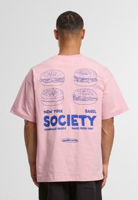 Pink cotton t-shirt with short sleeves. Back features blue illustrations of bagels and text: "New York Bagel Society, Handrolled Bagels, Baked Fresh Daily."