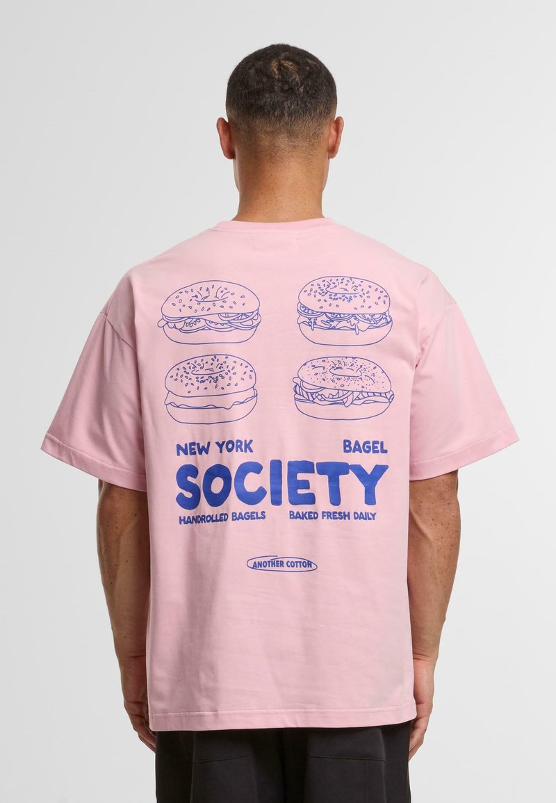 Pink cotton t-shirt with short sleeves. Back features blue illustrations of bagels and text: "New York Bagel Society, Handrolled Bagels, Baked Fresh Daily."