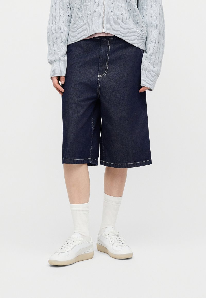 Personne portant un short en jean foncé jusqu'aux genoux, des baskets blanches, des chaussettes blanches et un pull gris clair en maille torsadée avec une fermeture éclair.