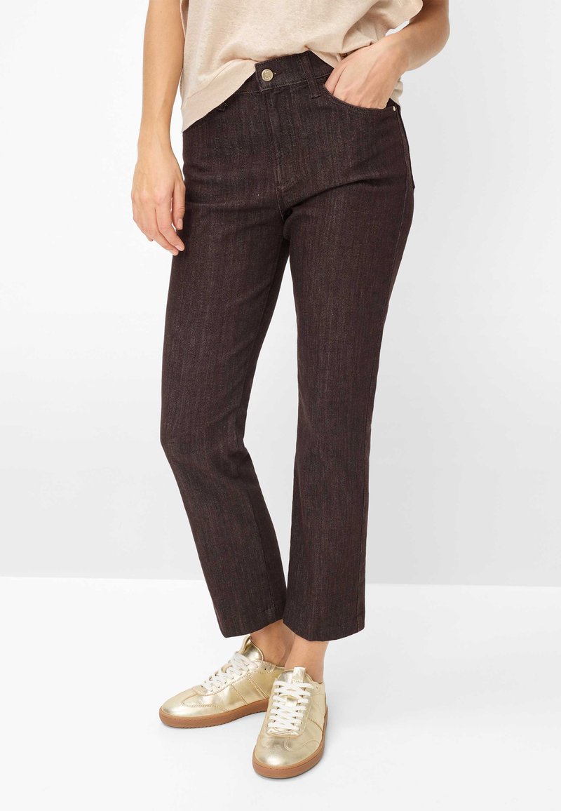 Person trägt dunkelbraune Jeans mit geradem Bein, beigefarbenes Oberteil und metallisch goldene Sneaker und steht mit einer Hand in der Tasche.