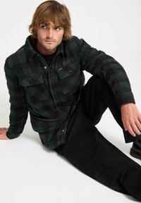 Green and black checkered flannel shirt with two front pockets, paired with black pants. Smooth fabric texture, relaxed fit.