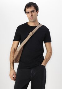 Bolso cruzado de gamuza beige con una correa ajustable de color marrón claro, que presenta una textura suave y un diseño simple y minimalista. Camiseta negra llevada debajo.