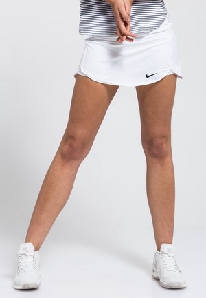 Jupe de tennis blanche avec détail froncé sur le côté, fabriquée en tissu léger. Portée avec un haut rayé et des chaussures de sport blanches.