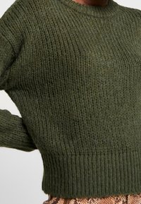 Grüner Strickpullover mit gerippter Textur, Rundhalsausschnitt und langen Ärmeln; verkürzte Passform und dicker Bund. Ideal zum Layering.