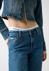 Τζιν μπουφάν και τζιν παντελόνι σε μεσαία μπλε απόχρωση, με κοντή γραμμή, κουμπιά με τρουκς μπροστά και λευκή ζώνη μέσης με την επιγραφή "GUESS JEANS".