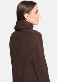 Femme aux cheveux bruns lisses regardant vers la droite, portant un pull à col roulé en tricot brun foncé sur un fond blanc.