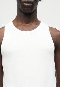 Ενήλικας που φοράει λευκό ριμπ αμάνικο τοπ, με εμφανές λαιμό, ώμους και άνω μέρος στήθους σε απλό φωτεινό φόντο.