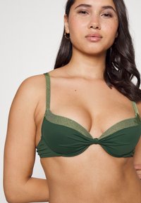 Dunkelgrüner Bikini-Oberteil mit einem weichen, strukturierten Band, das ein verdrehtes Vorderdesign aufweist und kontrastierende, hellgrüne Kanten entlang der Ränder besitzt.