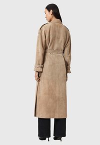 Μπεζ μακρύ παλτό από σουέτ με ζώνη στη μέση, πλαϊνές σχισμές και λεπτομέρειες στα μανίκια. Φοριέται πάνω από σκούρο παντελόνι με φαρδιά μπατζάκια και μπότες με τακούνι.