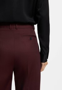 Pantaloni tailored color bordeaux realizzati in tessuto morbido, con una tasca posteriore con bottone nero e pieghe stirate, abbinati a una blusa nera.