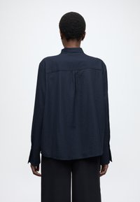 Dlhá košeľa námorníckej modrej farby s textúrou, voľného strihu, so zapínacími manžetami a záševkom na chrbte. Nosí sa s čiernymi nohavicami s širokými nohavicami.