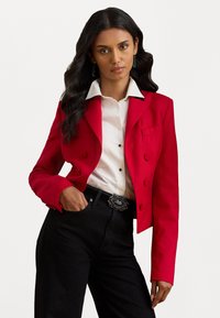 Červený blejzer s dvojitými gombíkmi, špicatými klopami a priliehavým strihom, skombinovaný s bielou košeľou a čiernymi džínsami, s dekoratívnou sponou na opasku.