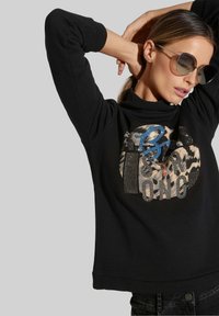 Schwarzer Pullover mit hohem Kragen, versehen mit einem gemusterten Grafikdesign in Beige, Blau und Strasssteinen. Glatte Textur, lange Ärmel.