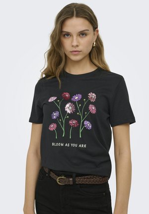Giovane donna che indossa una t-shirt nera con fiori colorati e testo "Bloom as you are,", abbinata a pantaloni neri e cintura intrecciata marrone.