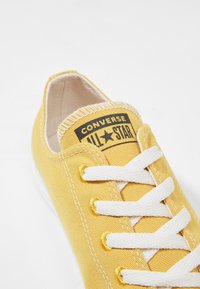 Baskets en toile jaune avec lacets blancs et coutures, arborant une étiquette noire "Converse All Star" sur la languette et une forme à bout arrondi.