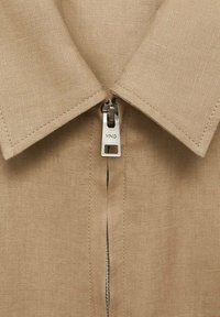 Beige skjorte med krave og metalzipper samt label, lavet af tekstureret stof, som viser et rent og simpelt design med synlige syninger.