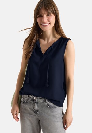 Mujer sonriente con blusa azul marino sin mangas con lazos de borlas y vaqueros gris claro, de pie frente a un fondo blanco.