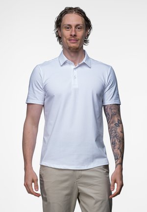 Jonge man met krullend haar draagt een wit poloshirt en beige broek, met een gedetailleerde tatoeagesleeve op zijn rechterarm.