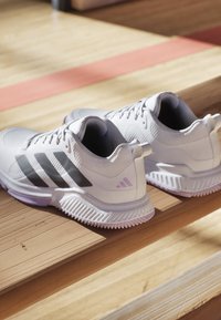 White athletic shoes with mesh upper, black stripes, and lavender accents. Textured sole with ridges and a pull tab at the heel.
