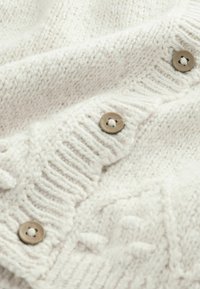 Hvid strikket cardigan med et tekstureret mønster og fire runde brune knapper med metallisk finish. Blødt, hyggeligt materiale.