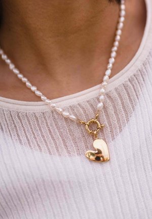 Perlenkette mit einem goldenen, herzförmigen Anhänger, getragen über einem weißen, halbtransparenten, gerippten Oberteil am Hals und Brust einer Person.