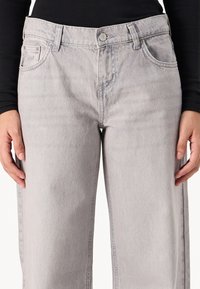 Lys grå denim jeans med rett passform, fem lommer og knappelukking. Slitt tekstur nær lommene gir en subtil detalj.