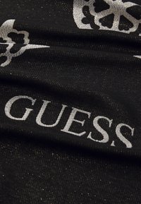 Mustmust kangas peene sära, millel on suured hõbedased tähed, mis kirjutavad "GUESS" ja dekoratiivsed lillemustrid.