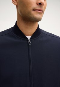 Zbliżenie na mężczyznę w ciemnogranatowej kurtce zapinanej na zamek z prążkowanym kołnierzem, noszonej na białej koszuli.