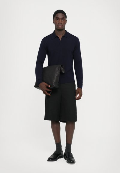 Marinblå långärmad piké, svarta shorts och svart läderväska. Modellen har på sig grå strumpor och svarta skor. Enkel design, inga synliga mönster.