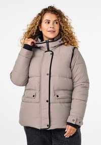 Giacca puffer beige con cappuccio, design imbottito, accenti neri, chiusura con zip e due tasche frontali. La texture morbida e liscia aumenta il calore.