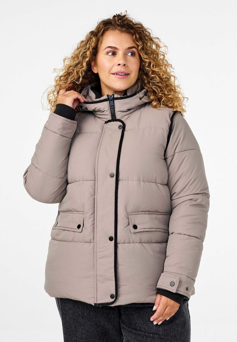 Giacca puffer beige con cappuccio, design imbottito, accenti neri, chiusura con zip e due tasche frontali. La texture morbida e liscia aumenta il calore.