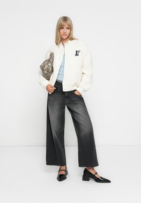 Grosse cardigan blanche avec un logo, haut bleu clair, jeans noirs larges et chaussures à talons noires, portant un sac gris à motif serpent.