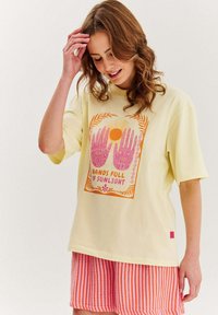 Světlé žluté oversized tričko s vícebarevným grafikou znázorňující ruce a slunce, doplněné oranžovými a bílými pruhovanými kraťasy.