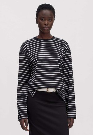 Vrouw draagt een zwart-wit horizontaal gestreept shirt met lange mouwen, ingestopt in een zwarte rok met een witte riem.