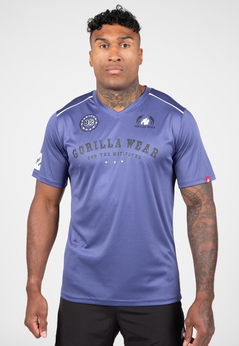 Paarse atletische shirt met V-hals en korte mouwen, voorzien van de tekst "GORILLA WEAR", logo en patches. Glad materiaal met contrasterende schouderaccenten.