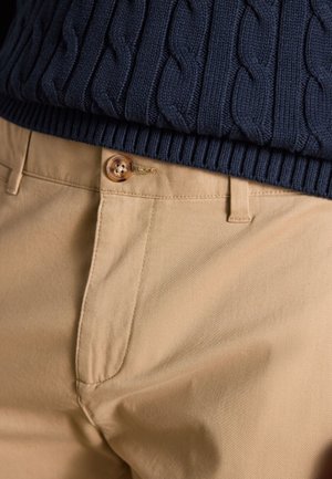 Nahaufnahme von beigen Hosen mit Knopfverschluss kombiniert mit dem Saum eines marineblauen, kabelgestrickten Pullovers.