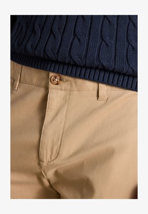 Nahaufnahme von beigen Hosen mit Knopfverschluss kombiniert mit dem Saum eines marineblauen, kabelgestrickten Pullovers.
