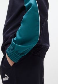 Νάυλον μπλε τοπ φόρμας με τιρκουάζ σατέν διακοσμήσεις και λευκές ραφές· διαθέτει ελαστικά μανσέτες και άνετη εφαρμογή· λογότυπο PUMA μπροστά.