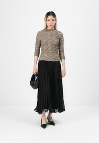 Jupe plissée noire longue associée à un haut texturé marron. Le modèle tient un sac noir et porte des escarpins noirs à bouts pointus. Fond simple.
