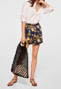 Nainen, joka on pukeutunut valkoiseen nappipaitaan, nauhakuosiset tummansiniset shortsit ja ruskeat sandaalit, pitää mustaa verkokassia, jonka sisällä on beige esine.