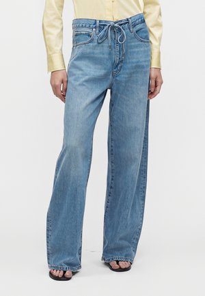 Personne portant un jean bleu clair à jambes larges avec une taille à cordon, une chemise jaune pâle à manches longues et des sandales noires à orteils ouverts.