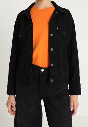 Personne portant une chemise orange vif sous une veste en jean noire boutonnée et un pantalon noir taille haute sur un fond uni.