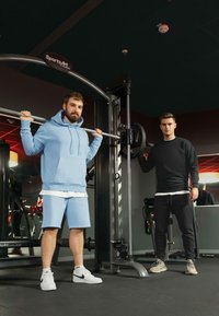 Twee mannen in een sportschool. Eén in een lichtblauwe hoodie en shorts, gewichten aan het tillen; de ander in een zwart sweatshirt, helpt.