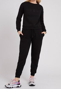Felpa nera con logo in rilievo, abbinata a pantaloni tapered coordinati. Il modello indossa sneaker colorate con una miscela di texture e tonalità.