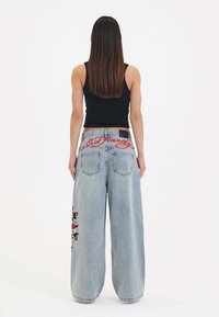 Světle modré široké džíny s vyšitým designem na levé noze a červeným logem "Ed Hardy" na zadní straně. Mají vysoký pas a denimovou strukturu.